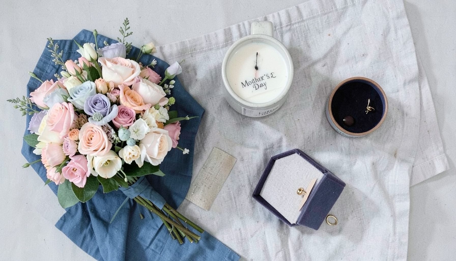 Overhead flat-lay of Mother's Day gifts: a pastel bouquet, a ceramic mug with a tea tag, a scented candle, and a small jewelry box on a linen cloth.