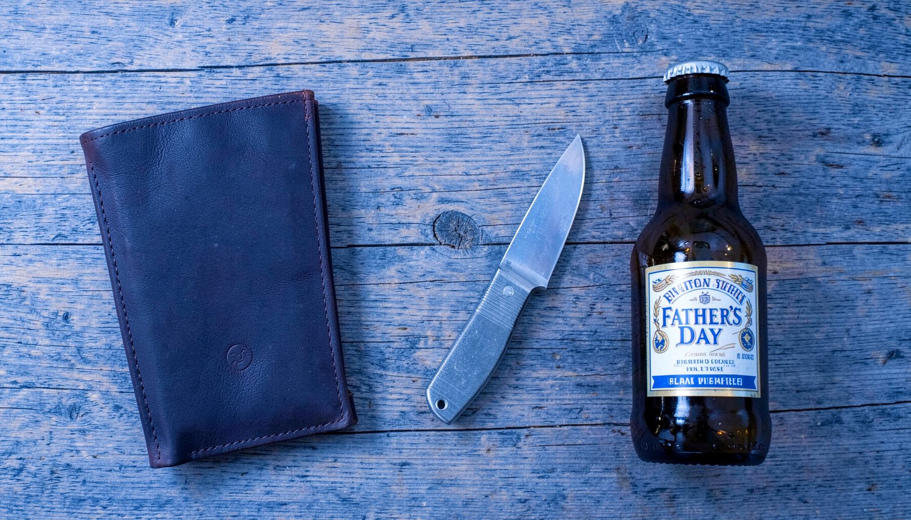 Overhead flat-lay of Father's Day gifts: a dark leather wallet, a stainless steel watch, a pocket knife, and a craft beer bottle on a rustic wooden table.