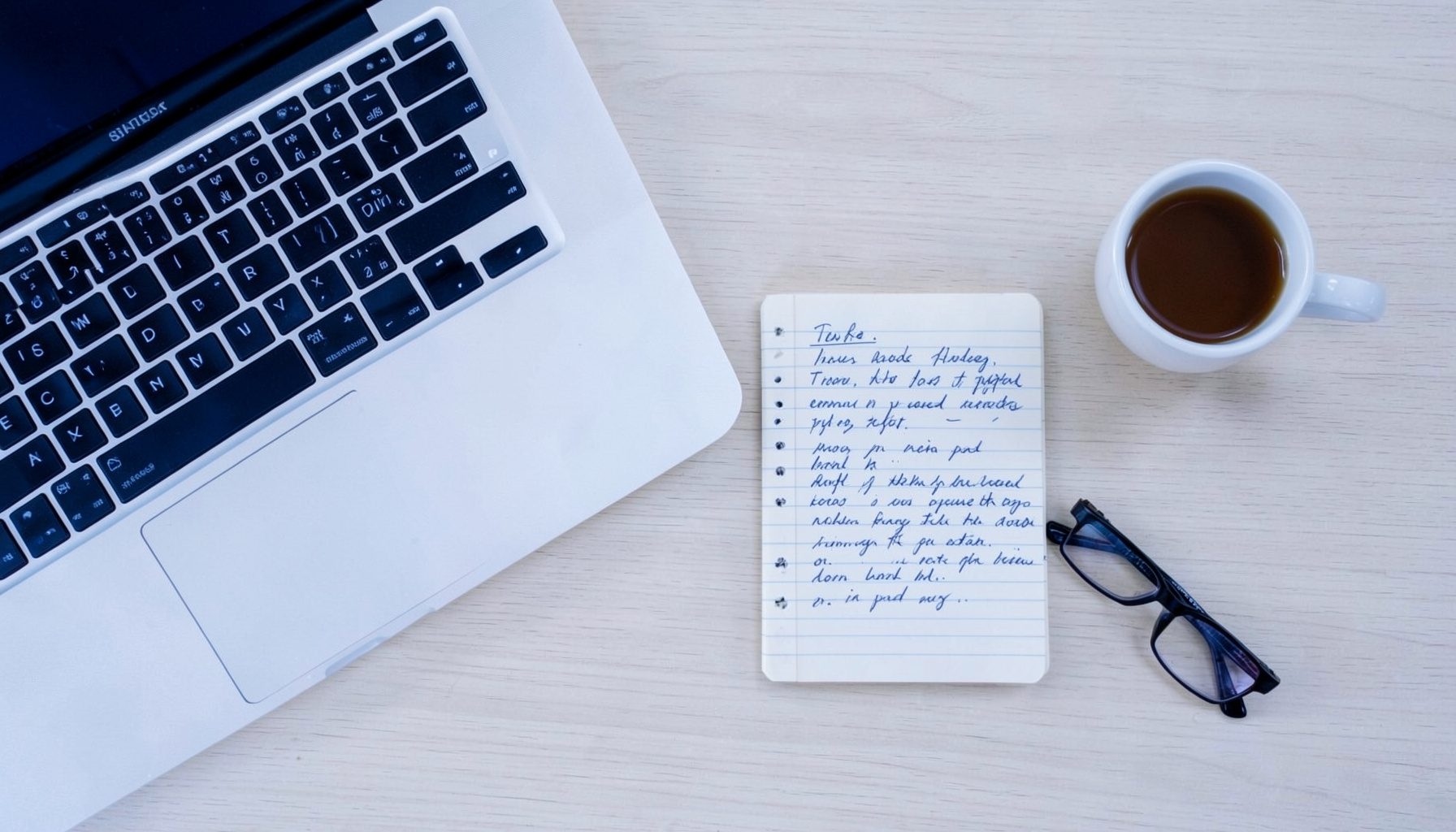 Overhead flat-lay of a blogger's desk: a laptop, a notebook with handwritten notes, a cup of coffee, and a pair of glasses on a light wood desk.