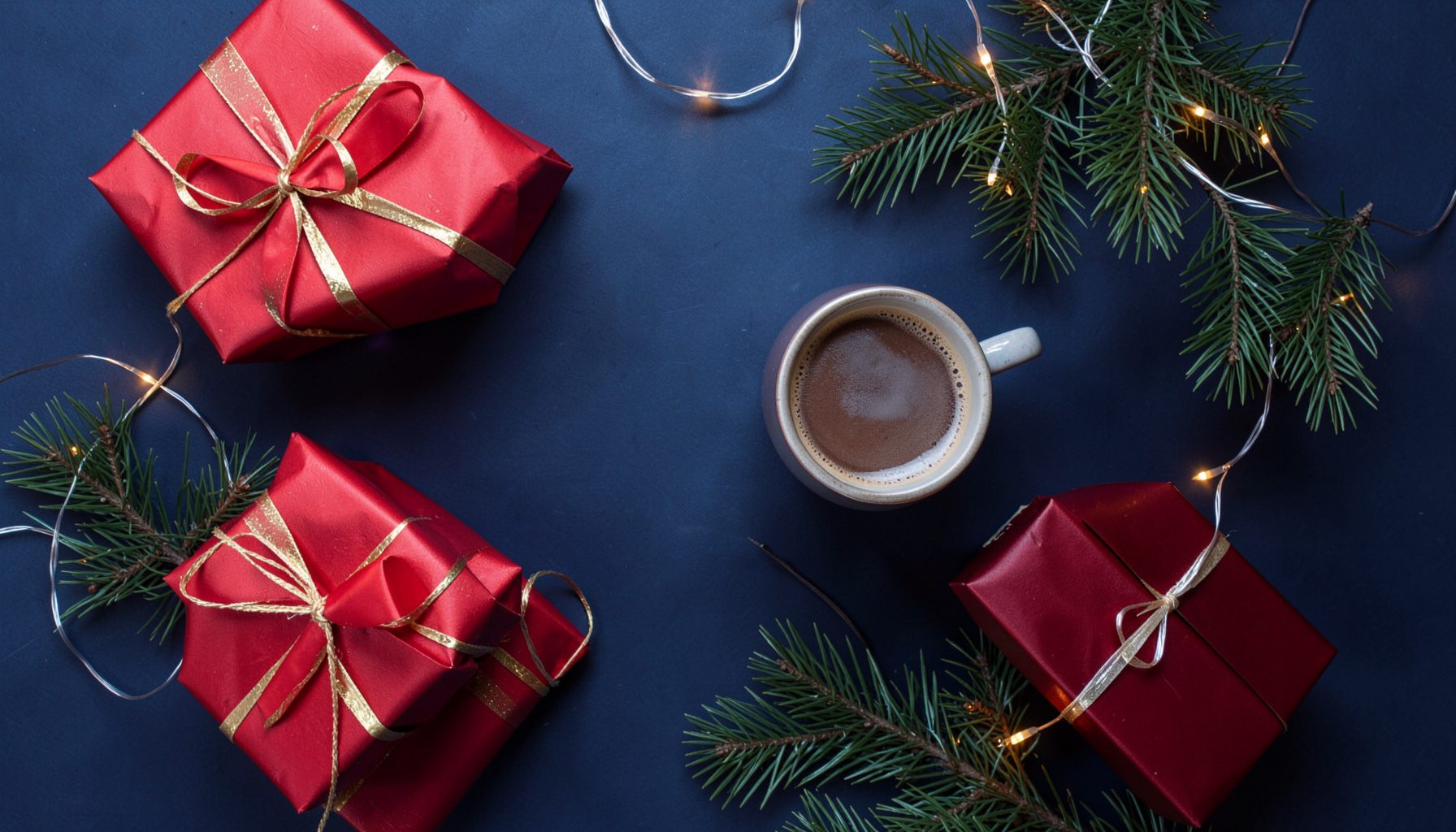Overhead flat-lay of holiday gifts: wrapped presents in red and gold paper, pine branches, string lights, and a steaming mug of cocoa on a dark surface.
