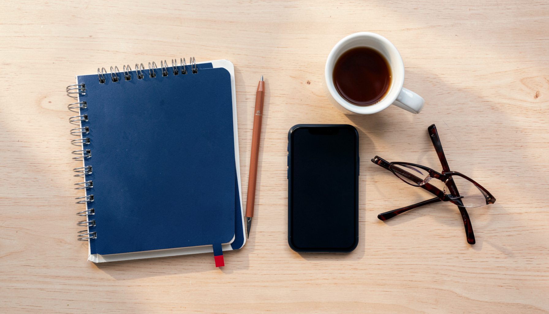 Overhead flat-lay photograph of lifestyle blogging tools: a notebook, a smartphone, a cup of coffee, and a pair of glasses arranged neatly on a light wooden desk surface, with warm natural light and soft shadows.