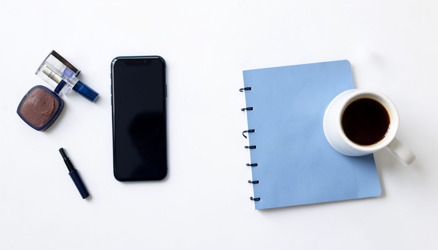 Overhead flat-lay photograph of lifestyle products on a bright white surface: a smartphone, beauty items, a notebook, and a coffee cup arranged with generous negative space, conveying a clean editorial mood